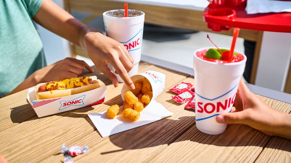 Friends sharing a SONIC chili cheese coney, tater tots and cherry limeade at an outdoor table